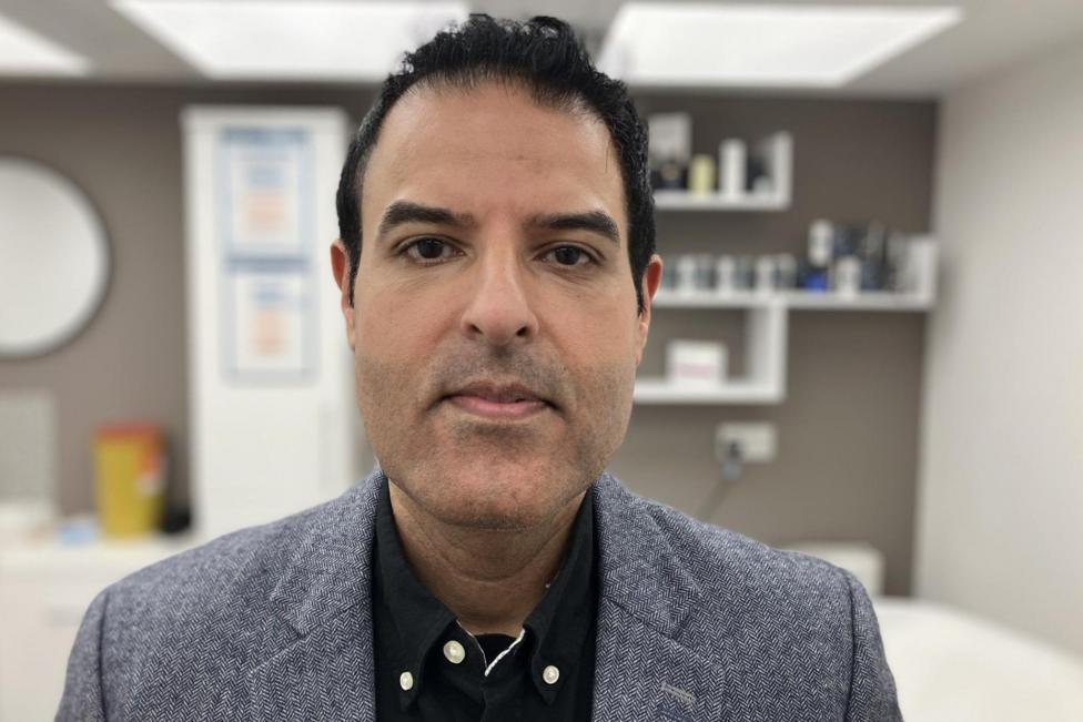 The image is a close-up portrait of a person positioned centrally in an indoor setting. The person is wearing a textured grey blazer over a dark button-up shirt and is facing directly toward the camera. The background is softly blurred and appears to be a clean, modern office, clinic, or consultation room with shelves, framed items, and various small objects visible.