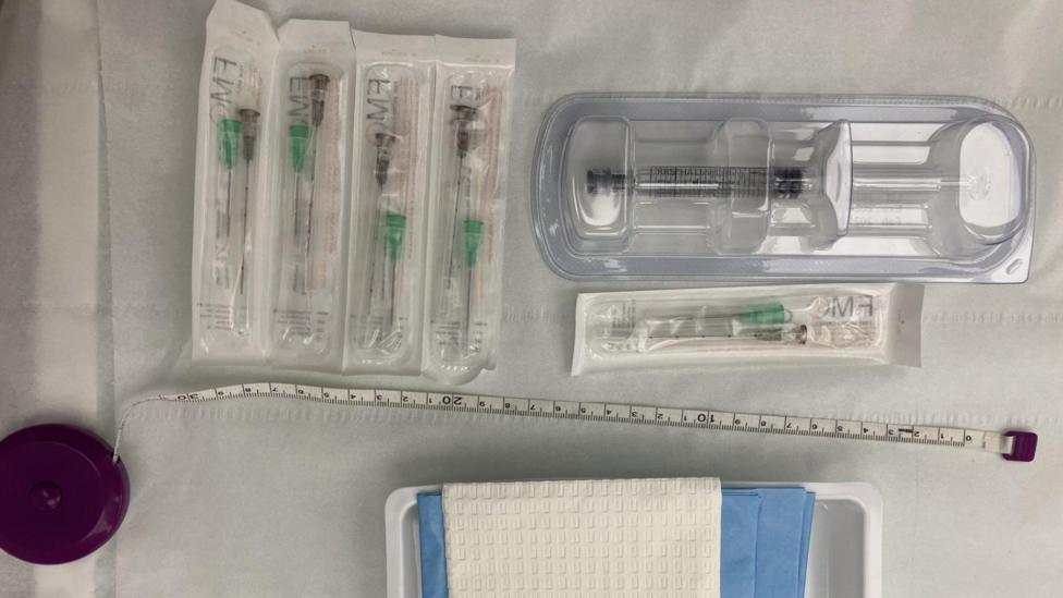 The image shows a neatly arranged set of medical supplies laid out on a sterile-looking white surface. Several individually packaged cannulas are positioned on the left, while a larger pre-packaged syringe sits in a clear plastic tray on the right. A measuring tape stretches horizontally across the middle of the image, and a tray containing absorbent pads or dressings is visible near the bottom edge.