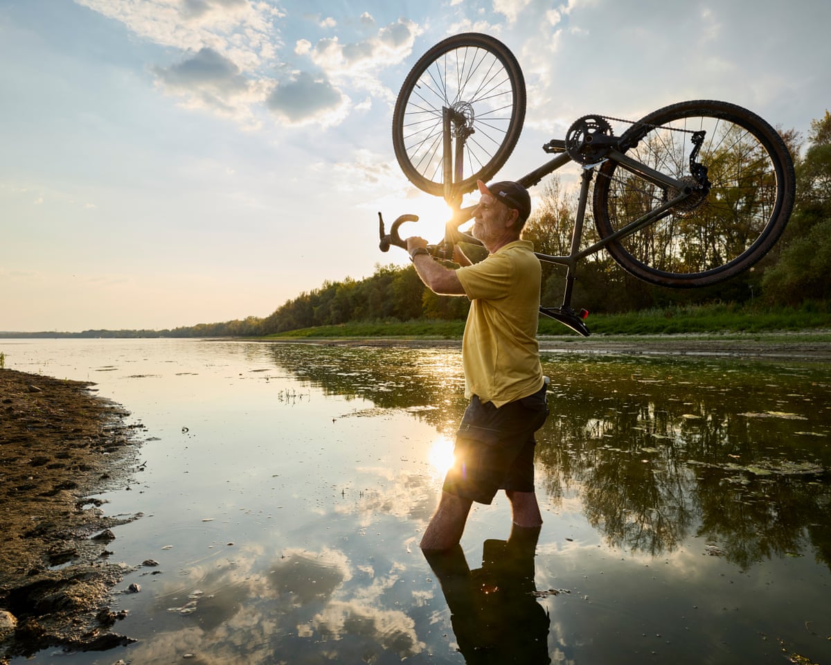 Kürti wades through water, carrying his bike on one shoulder
