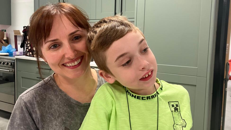 A woman with short brown hair smiles at the camera in a grey-tshirt, while her son, who is on her left, sitting on her knee looks slightly of camera to the right as he wears a bright yellow t-shirt. They sit in a kitchen with green panelling and a white floor. 