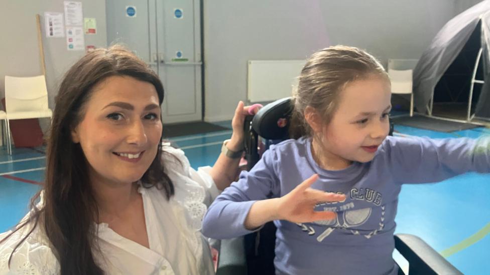 In a sports hall with a blue floor a woman in a white top smiles to the camera as she has her hand on the back of her daughters wheelchair. The woman, who is kneeling beside her daughter is on the left of her daughter. Her daughter looks of to the right of the camera as she sits wearing a lilac top. 