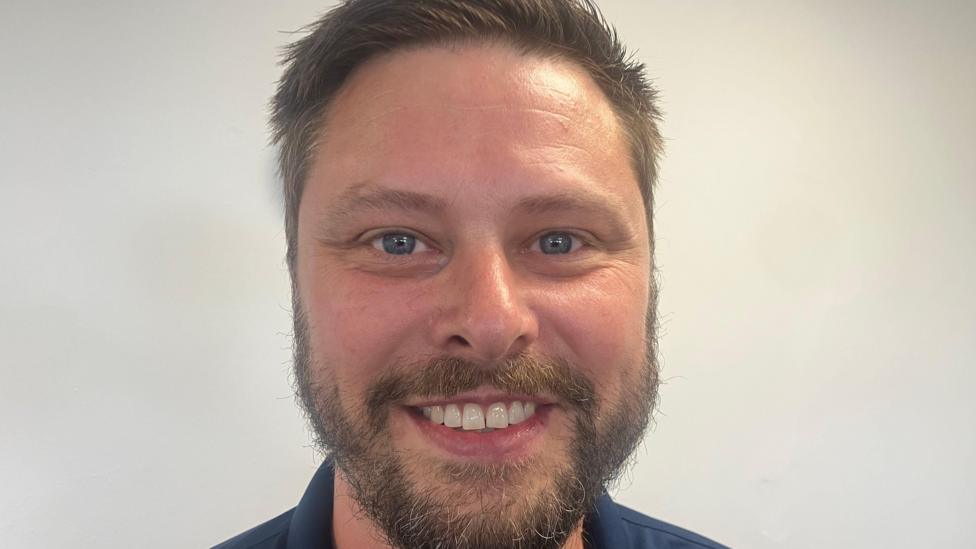 A man with short brown hair and a short brown beard and moustache smiles to the camera. 
