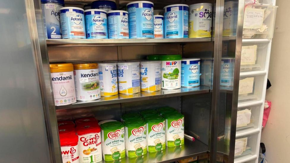 Three metal shelves are lined with colourful tubs of baby milk. There is a tall, white set of drawers on the right with paper taped on each drawer denoting what is in it.