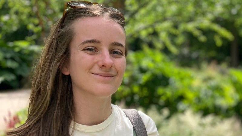 Close up of Bristol University researcher who is smiling at the camera and stood in front of green bushes outside.