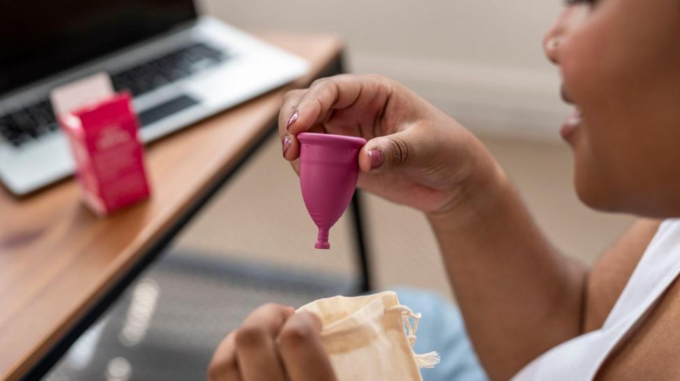 A woman to the right holding a pink menstrual cup in her hand. She is holding a small fabric pouch in her other hand. In the background a opened box can be seen on a table, stood next to an open laptop.