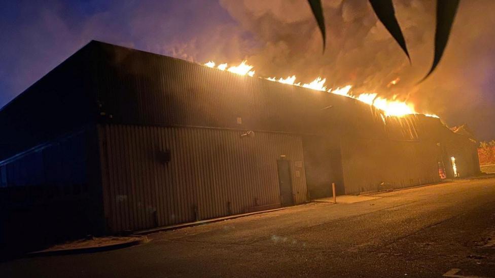 A warehouse on fire in Harwich. The fire service responded to the incident in the early hours of the morning therefore the picture has been taken in the dark, however, the warehouse is lit up by flames. A plume of smoke coming off the building is stretching into the sky. Where there are flames on the right hand side of the building in the picture, the roof has started to warp.