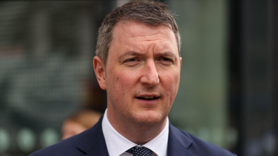 John Finucane. He has short brown hair, wearing a navy blazer, white shirt and navy polka dot tie.