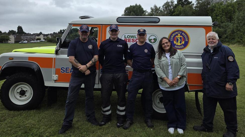A large 4-4 that is painted white, grey and orange is parked on grass. In front of it is five people who are smiling at the camera. There are four men and one woman. The men are wearing all navy outfits with a logo on the chest. The woman has dark hair and is wearing a pale green jacket and white t-shirt. 