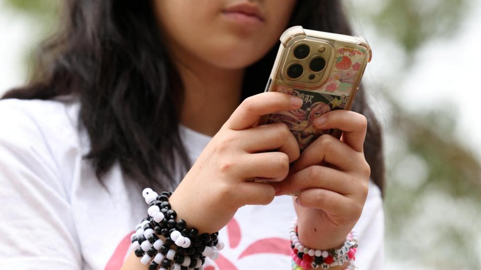 A teenage girl wearing coloured bracelets sits outside looking down at her iPhone