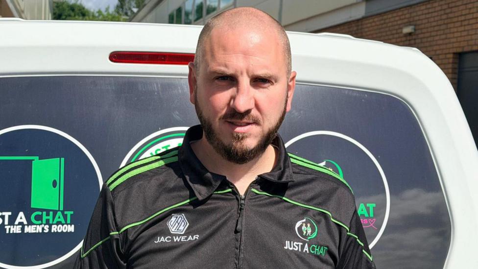 A man wearing a black and green "Just a Chat" training top stands in front of a branded white van outside a building.