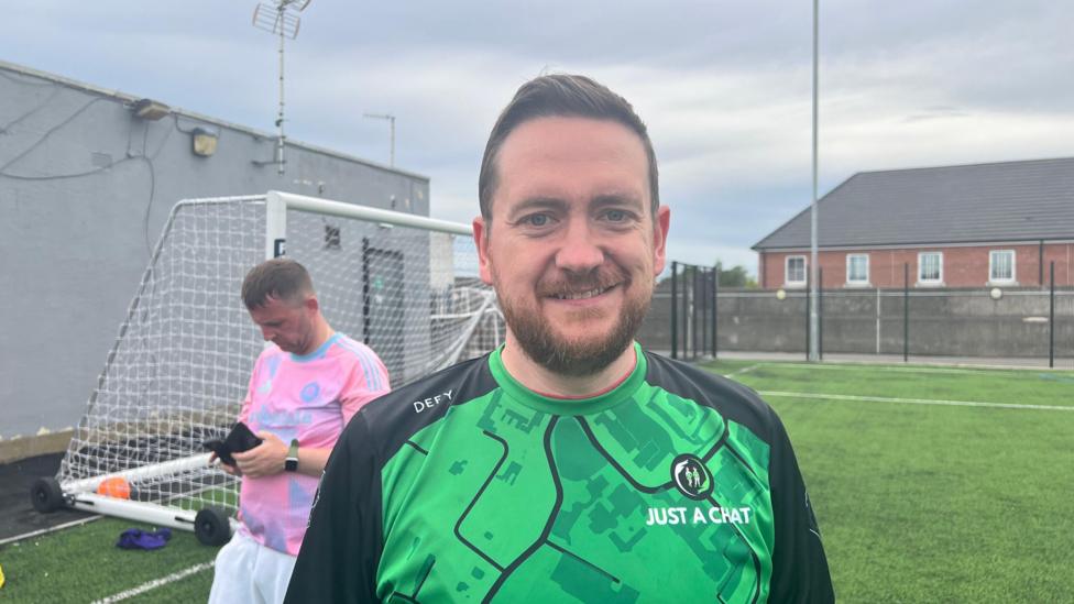 A smiling man is wearing a green and black "Just a Chat" football jersey stands on an outdoor football pitch. Another player in a pink shirt is looking down at his phone near a goal post under an overcast sky