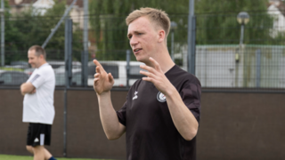 A man is wearing a black top and is coaching people on a football pitch.
