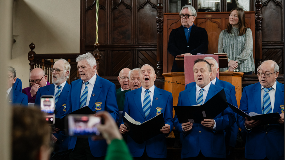 Aelodau Côr Meibion Pontypridd yn canu pan ailagorodd y capel ym mis Chwefror, gyda Colin Lewis (cadeirydd Capel Rhondda.Cyf) a Rhian Hopkins yn sefyll y tu iddyn nhw yn y sêt fawr