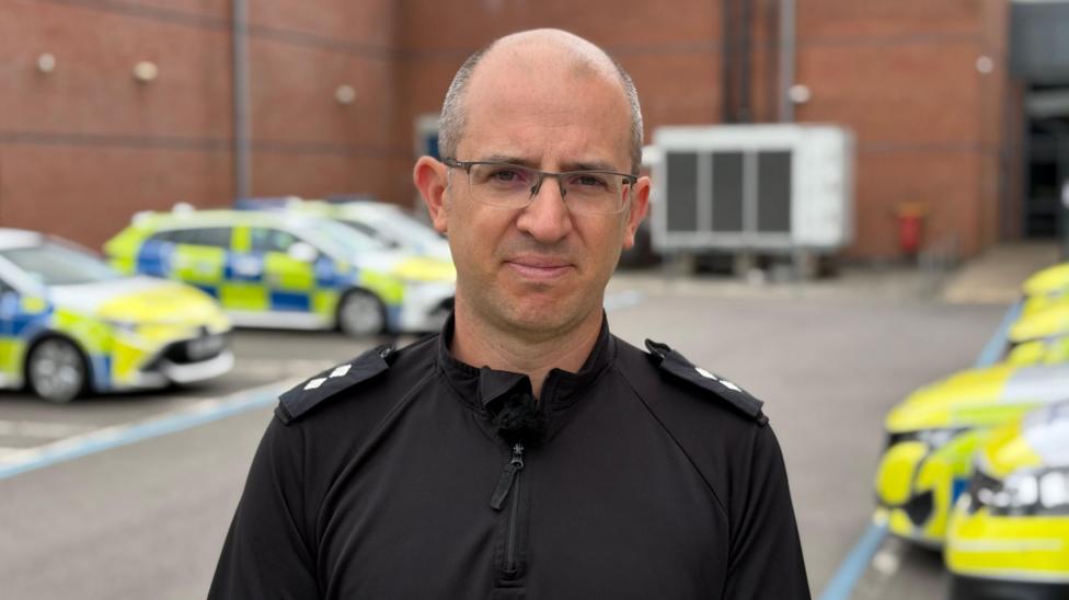 Inspector Joe Senior is pictured standing outside a police station. He is wearing a black police uniform and a pair of glasses. He has grey receding hair. There are several police cars parked in the station car park in the background.
