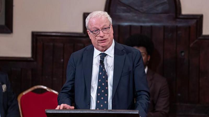 Lord Chris Smith wearing a navy blue suit and white shirt with matching navy blue tie with the Cambridge University crest. He has white hair and black rimmed glasses. He is standing behind a podium.