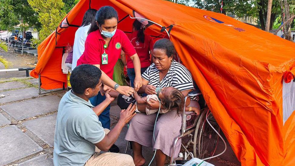 A woman sitting in a wheelchair outside a bright orange tent holds a small infant in her arms, an oxygen mask over its face, as a man and a woman tend to the child
