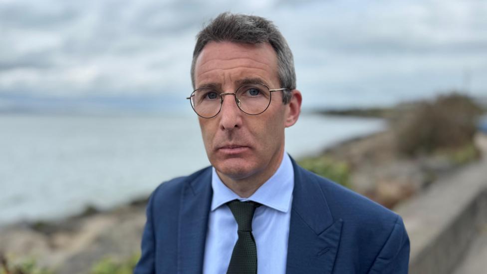 Andrew Muir, a man with greying hair and glasses, looks to the camera at a seafront setting. He is wearing a navy suit jacket a blue shirt and a black tie.