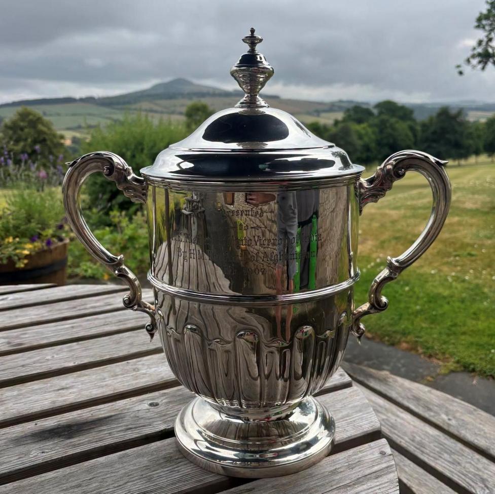 A silver trophy with two ornate handles sitting on a wooden table.