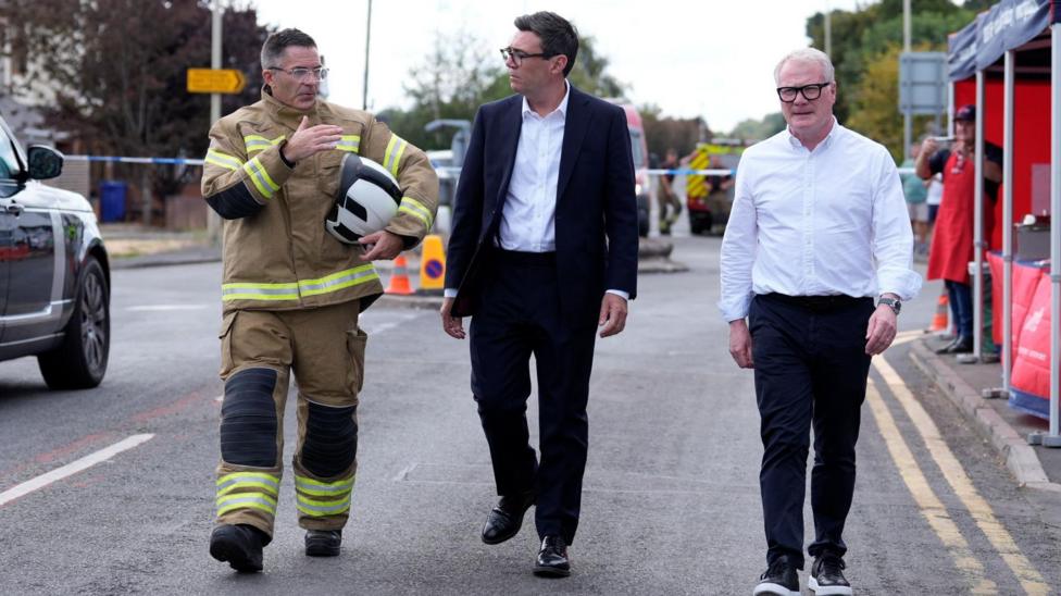 Andy Burnham, in a dark suit and white shirt, walks down a street alongside chief firefighter Simon Tuhill and the West Midlands mayor Richard Parker