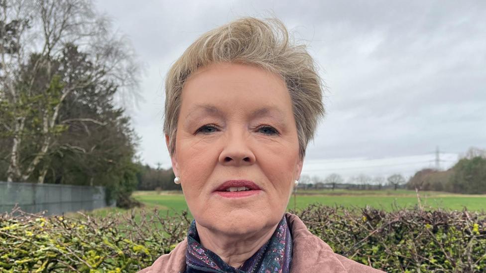 A woman with short fair hair standing in front of a hedge with fields in the background. She is wearing a light brown top and paisley scarf.