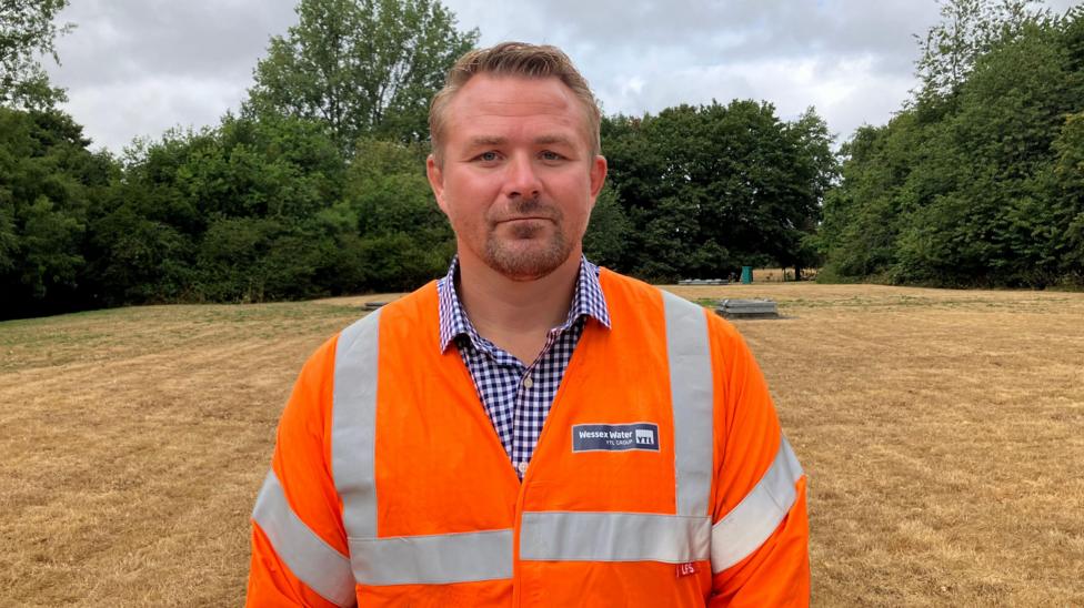 A man with short brown hair and a goatee beard looks at the camera, wearing a orange hi-vis jacket over a blue and white checked shift, and standing on a covered reservoir. There is brown grass and green bushes and trees in the background.
