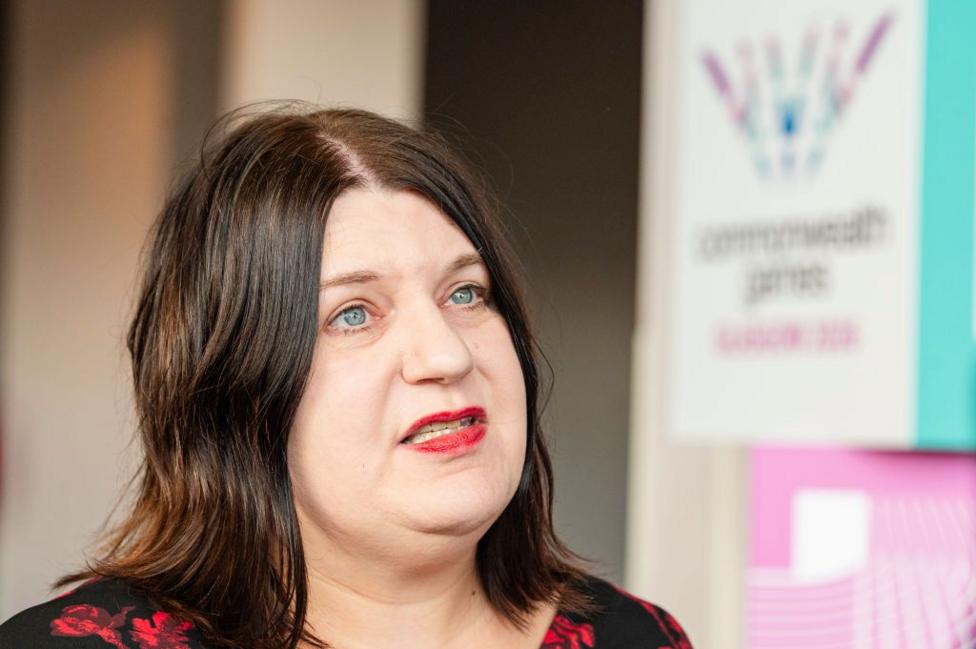 Susan Aitken, a woman with dark hair to her shoulders and a black and red dress on, speaking to someone standing off camera. She is looking slightly sideways.