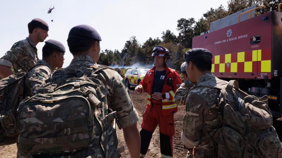 Soldiers from 10 Queen's Own Gurkha Logistic Regiment receive a briefing from a firefighter as a helicopter with water flies overhead