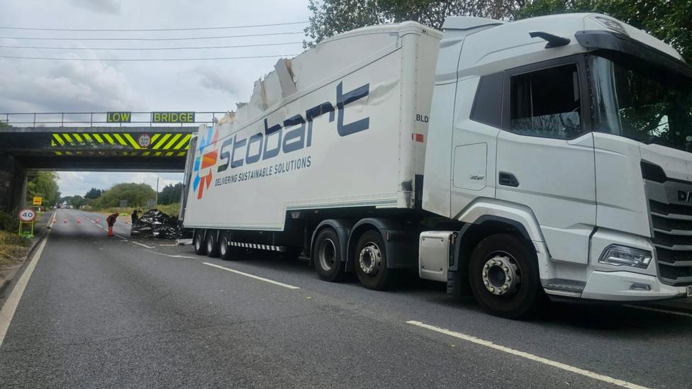 A road under a railway bridge is in the distance and at the front of the photograph is a lorry on the road with a damaged roof. There is debris and cones under the bridge.