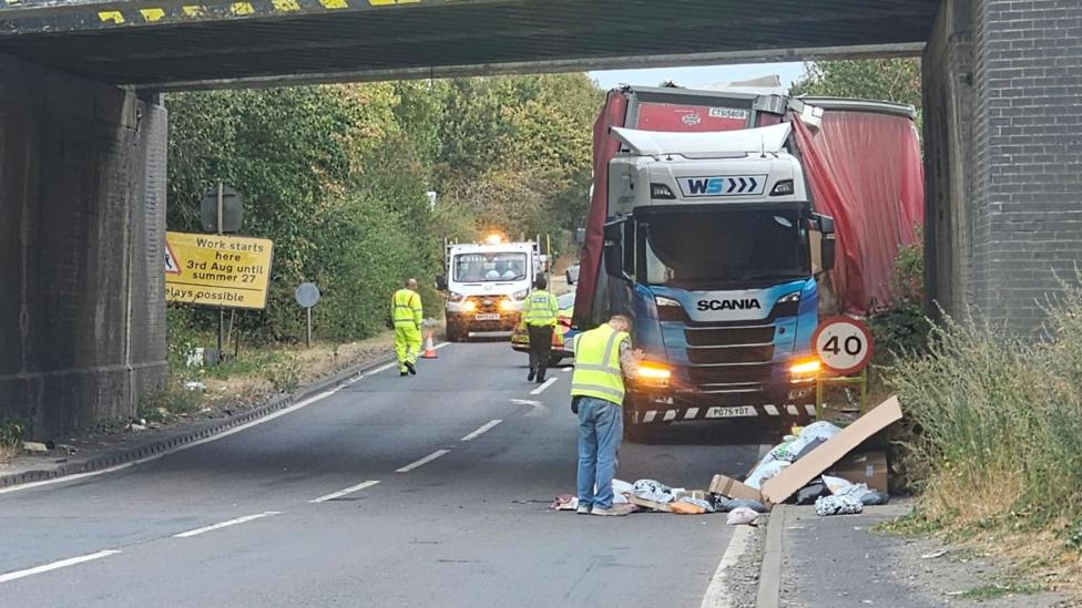 A road with a railway bridge over the top. A lorry is parked underneath the bridge and three people wearing hi-vi jackets are on the road and cones can be seen too.