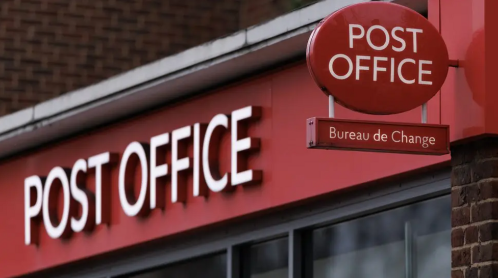 Close-up of a Post Office storefront with red branding, featuring large white “POST OFFICE” lettering across the shop fascia. A circular red Post Office sign and a smaller “Bureau de Change” sign are mounted above the entrance, with brickwork and windows visible in the background.