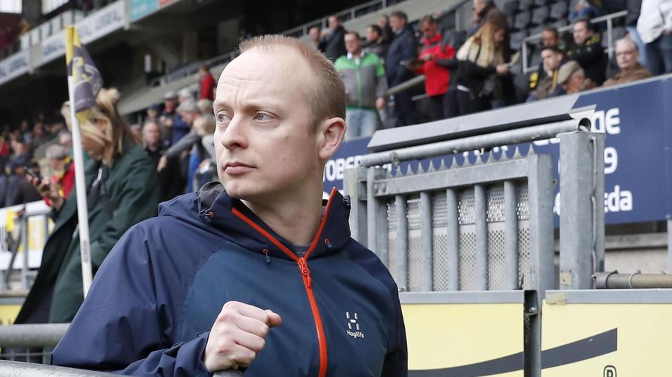 A man with short fair hair, wearing a blue zipped up jacket in a football stand.