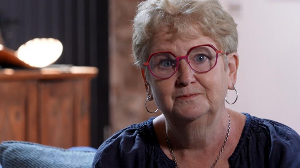 Dr Jane Dickson, a woman with short blonde hair and large red glasses, sits on a blue sofa, looking into the camera. She is wearing a dark blue top, silver hoop earrings and a silver necklace