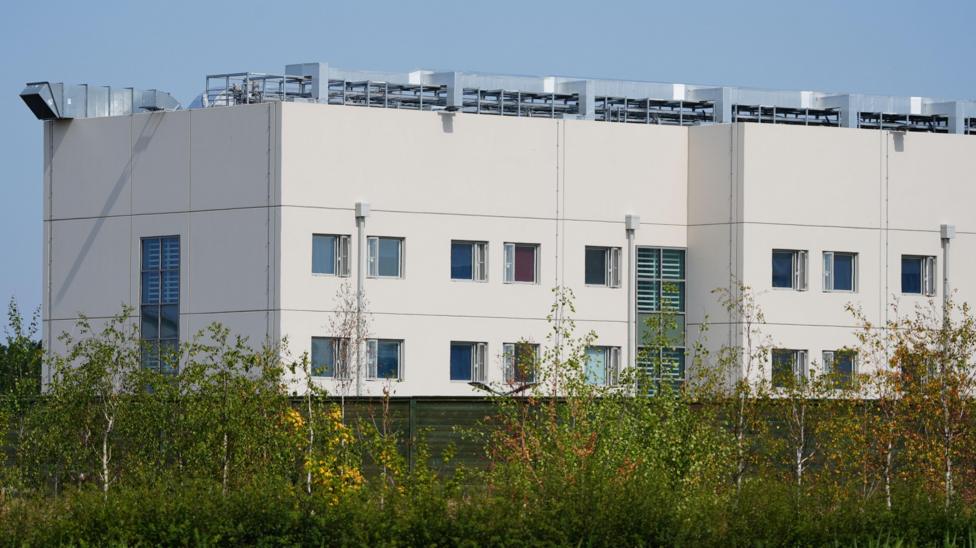 A modern-looking, white prison building. It appears to be four stories high with the central stories having two rows of windows. A ventilation system can be seen on the roof. Trees are growing in the foreground.