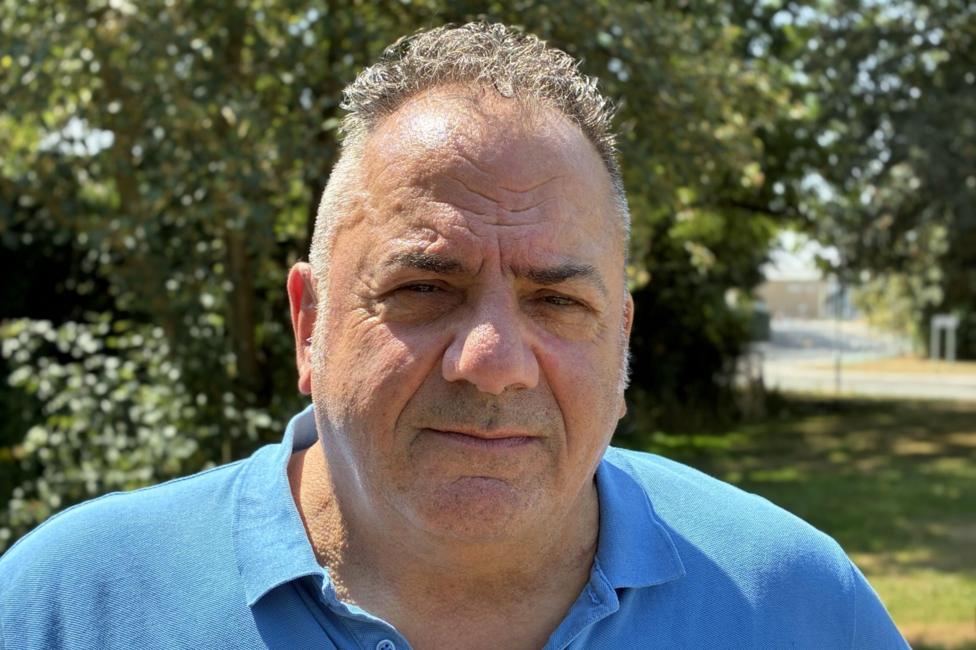 A middle-aged man with short cropped hair, curly on top, stands in sunlight in front of a patch of grass and green trees. He is wearing a blue polo shirt and has a serious expression on his face.