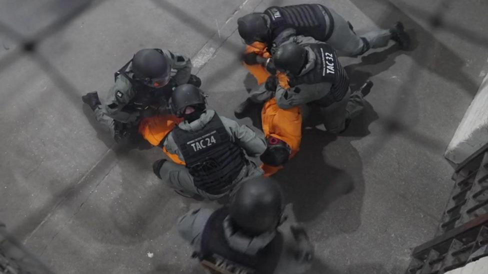 A view from above of five people, wearing black helmets, black stab vests and grey uniforms appearing to roll up orange fabric in a prison space with a dark grey concrete floor.