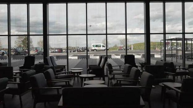 A view of what looks like a meeting lounge, with black upholstered chairs and small coffee tables, looking out through a bank of glass windows towards a car park.