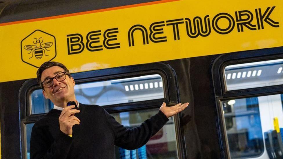 Andy Burnham, in a black jumper holding a microphone, stands in front of a yellow Bee Network vehicle