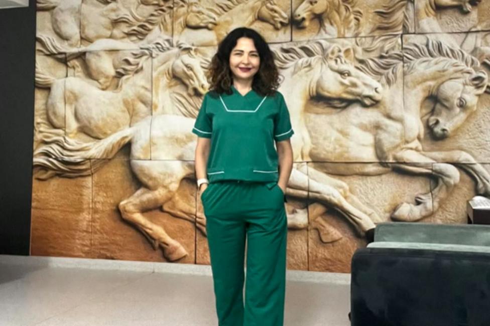 Firdevs in green medical scrubs stands indoors in front of a large sculpted wall mural depicting galloping horses in relief.