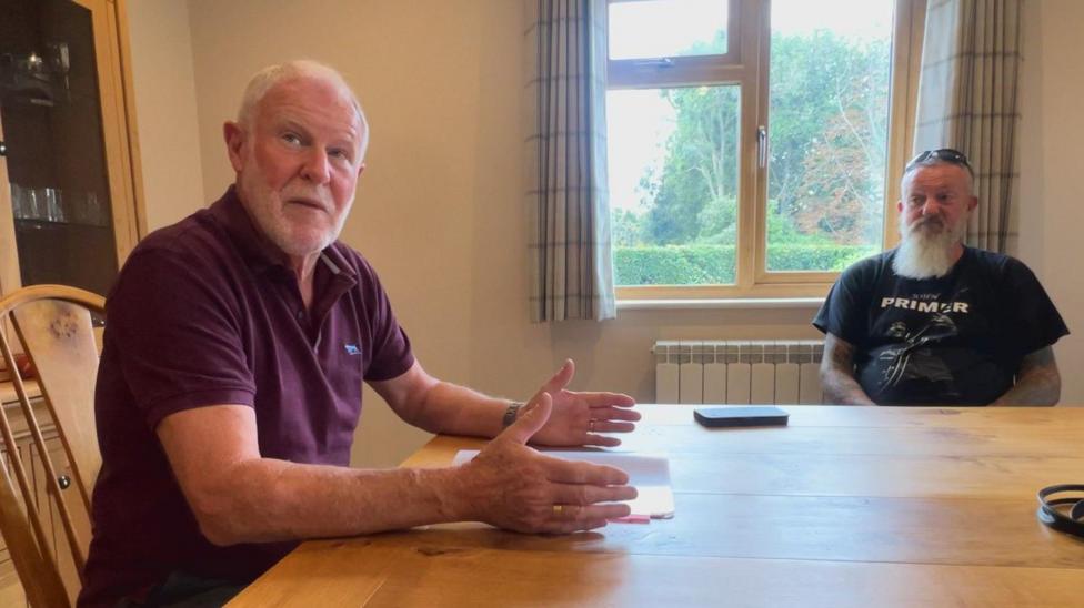 Two men sit at a wooden dining table. There is a document on the table and a phone. There is a window in the background showing greenery and trees.
