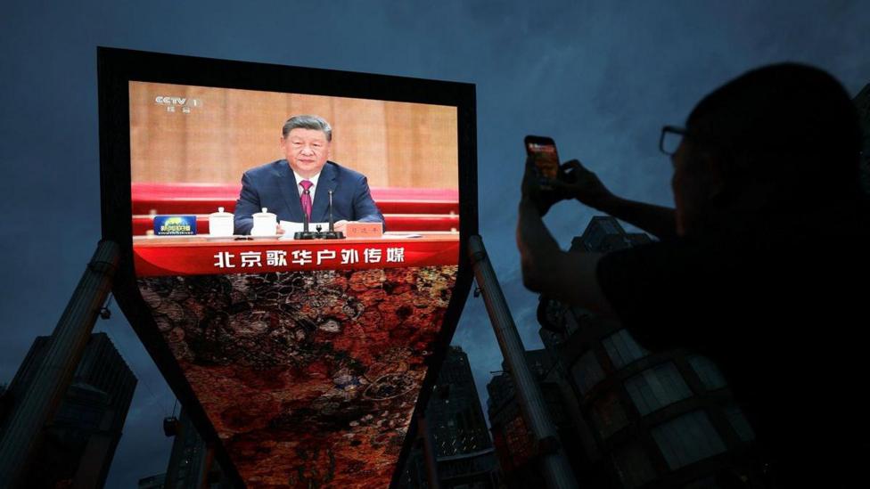 A person uses her mobile phone near a giant screen showing Chinese President Xi Jinping delivering a speech