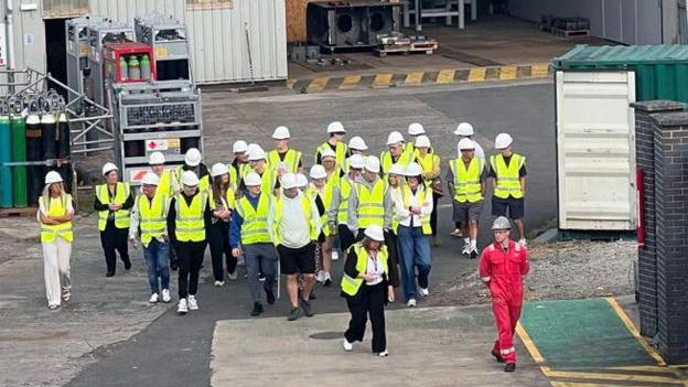A group of people in yellow high visibility vests are shown around an industrial site, led by a man in a red boilersuit