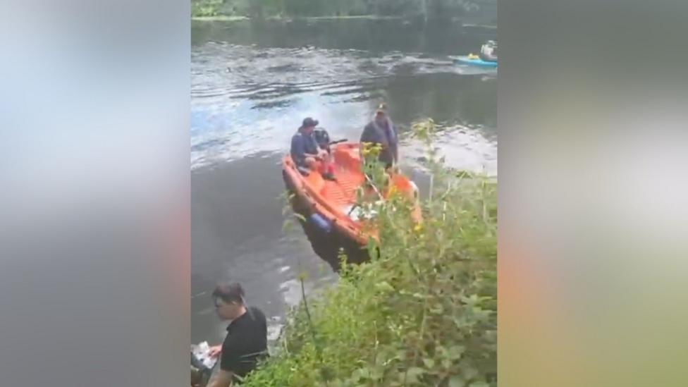 An orange rescue boat on the river Avon in Evesham. It has a man sitting in it, with another standing alongside. In the foreground is vegetation growing on the riverbank, while a man in a black T-shirt stands next to it in the water.