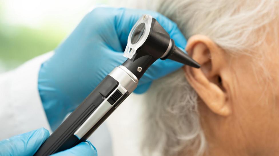 A close-up of a medical examination where a healthcare professional is inspecting a person's ear using an otoscope. The healthcare professional is wearing blue disposable gloves. The person being examined has short, grey hair. The background is softly blurred, keeping the focus on the ear and the otoscope.