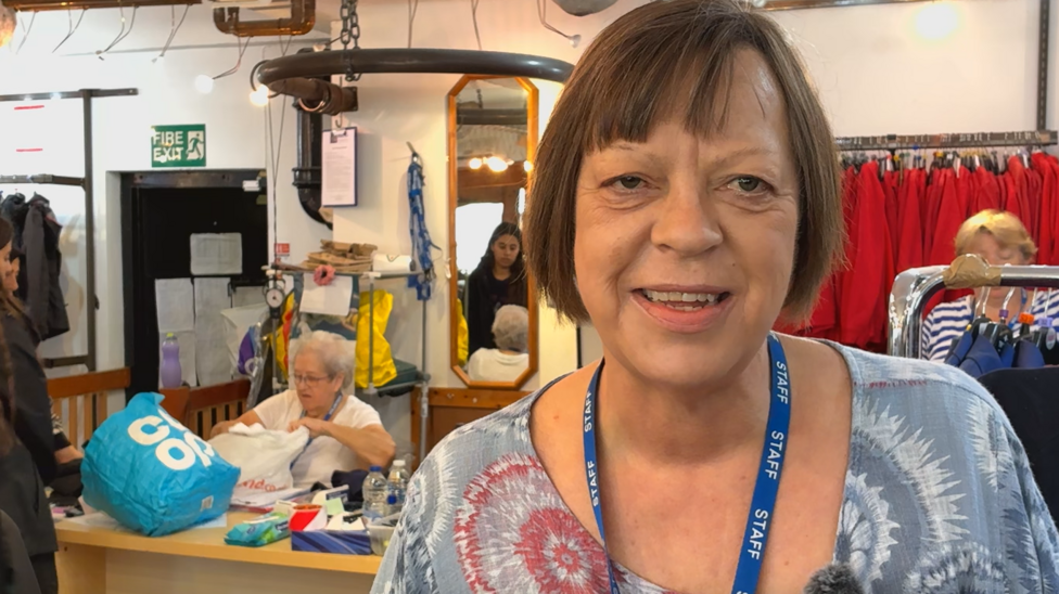 A middle-aged woman with brown hair cut into a bob, wearing a grey and white patterned blouse and a blue lanyard, smiles at the camera. Behind her are clothes rails while volunteers are sorting clothes and serving customers. 