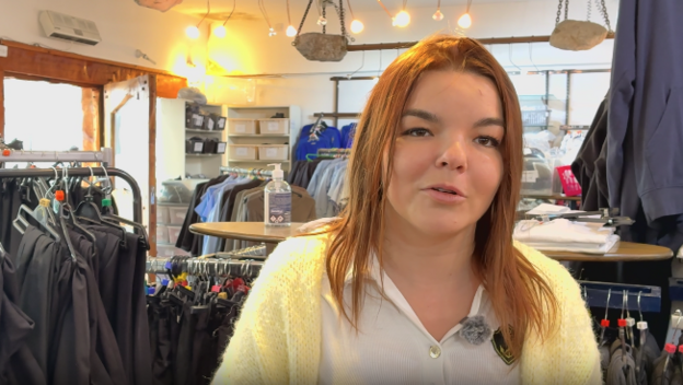 A young woman with long light brown hair, wearig a white shirt and a yellow woolen cardigan, speaks to the camera in front of racks of school uniform clothing.