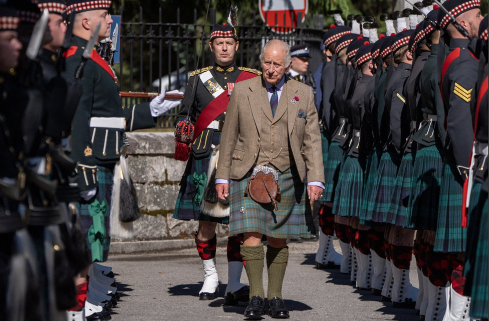 King Charles III, wearing a tweed jacket, waistcoat and tartan kilt, walks between two ranks of troops from Balaklava Company, 5th Battalion, The Royal Regiment of Scotland, during a ceremonial inspection. Soldiers in dark uniforms, tartan kilts and glengarry caps stand at attention on either side, creating a guard of honour along the roadway, while an officer in full ceremonial dress follows behind. The scene takes place outdoors in bright sunlight.