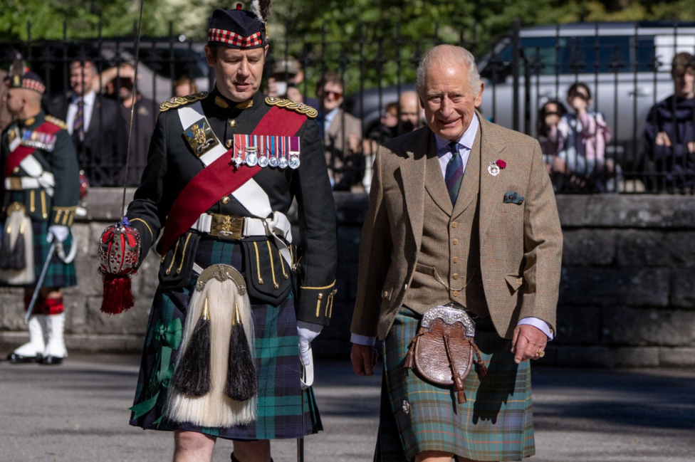 King Charles III walks alongside a soldier in ceremonial Highland dress during a parade inspection. The King is wearing a tweed jacket, waistcoat and tartan kilt, while the soldier wears a dark dress uniform decorated with medals, a tartan kilt and traditional military accessories. Spectators watch from behind railings in the background.