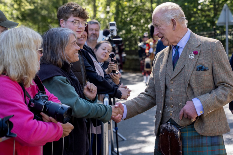 King Charles III greets members of the public gathered behind crowd barriers during a visit, shaking hands with one person while others look on and take photographs. The King is wearing a tweed jacket, waistcoat and tartan kilt. Several spectators hold cameras and smartphones as they line the roadside beneath trees in bright sunshine.