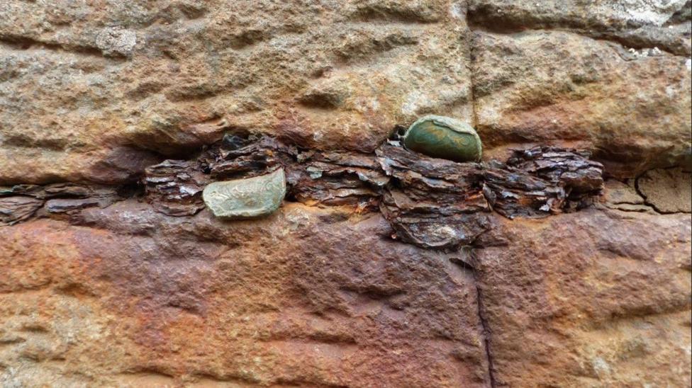 A line of old coins in the centre of the rock. The rock has turned a rusted orange colour around the coins. There is a large bulk of old rusted coins in the middle with some newer coins folded over.