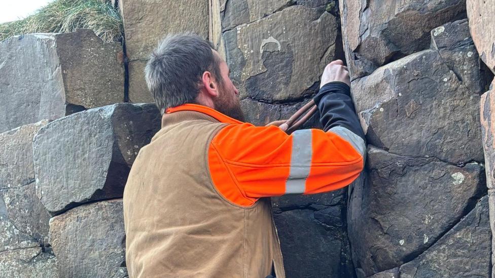 A man in work clothing uses told to remove coins from a rock. he is standing with his back to the camera